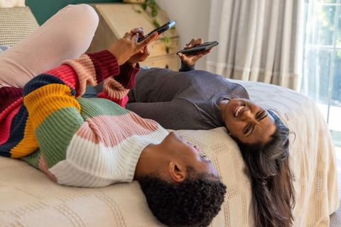 Couple lying on bed using smartphones and laughing in sunlit cozy bedroom