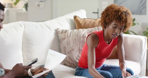 African american couple balancing bills on sofa, woman sorting invoices, man using phone