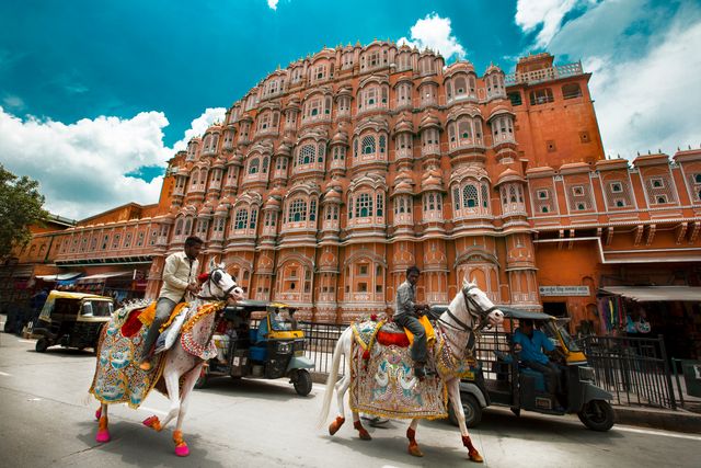 Colorful cultural parade passing in front of the elaborately designed Hawa Mahal, a historical palace in Jaipur, India. Decorated horses ridden by individuals in traditional attire underscore the rich cultural heritage and festive environment. Ideal for use in travel blogs, promotion of Indian tourism, cultural exhibitions, and educational materials about Indian architectural icons.