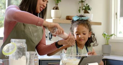 Mother and daughter baking together whisking batter pouring flour using tablet in sunlit kitchen