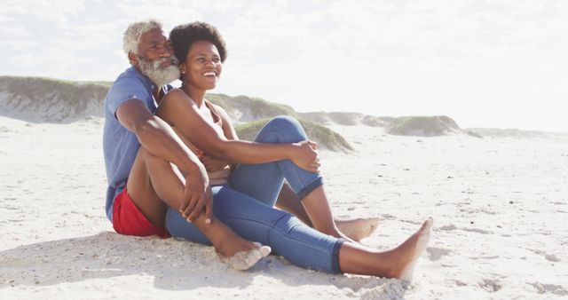 An African American senior couple enjoying a sunny day on the beach. They are sitting on the sand while smiling and looking content. There are dunes visible in the background. Ideal for use in retirement, lifestyle, outdoor leisure, and healthy living campaigns.
