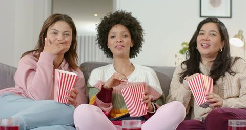 Diverse women enjoying cozy movie night on sofa with striped popcorn boxes and drinks