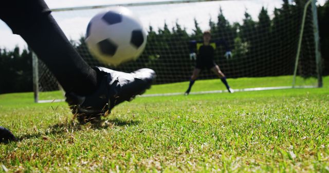 A soccer player in black cleats is kicking a ball towards the goal where a goalkeeper is ready to make a save, with copy space. Capturing the intensity of a soccer match, the focus is on the moment of impact and the anticipation of a goal.