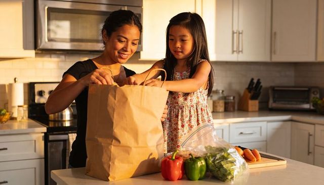 Sunlit mother and daughter unpacking groceries at modern kitchen island with fresh peppers