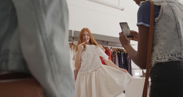 Friends enjoying time shopping in a clothing boutique. One friend showing off a white dress while another friend takes a photo with a smartphone. Perfect for use in fashion blogs, social media marketing, retail store promotions, or articles about trends and lifestyle.