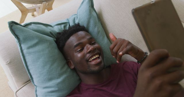 Man lying on sofa with cushions, smiling and engaging in video call on smartphone. Ideal for illustrating modern communication, relaxation at home, and technology use in daily life.
