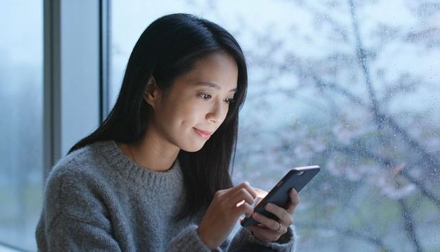 Asian woman using smartphone by rainy window in cozy gray sweater, contemplative mood