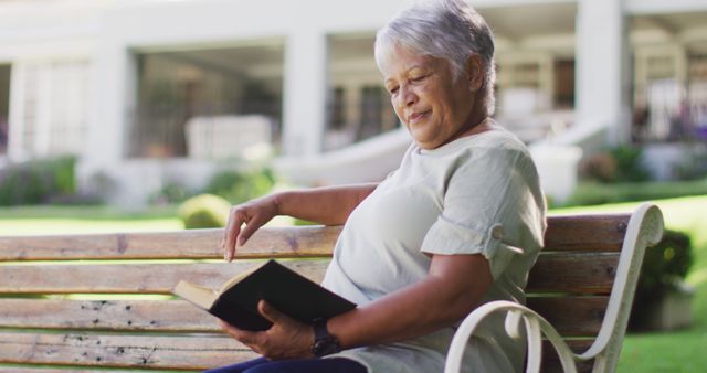 Image of relaxed biracial senior woman reading, sitting on bench in garden. active retirement lifestyle, hobby and spending time outdoors.