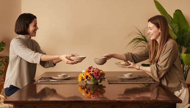 Two women passing bowls across dining table sharing cozy casual meal with flowers
