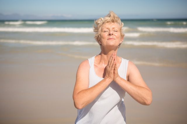Elderly woman stands on beach with hands clasped, embracing tranquility and mindfulness. Perfect for illustrating concepts of relaxation, mental wellness, elder care, and healthy lifestyles. Suitable for wellness blogs, senior living advertisements, meditation apps, and nature-themed backgrounds.