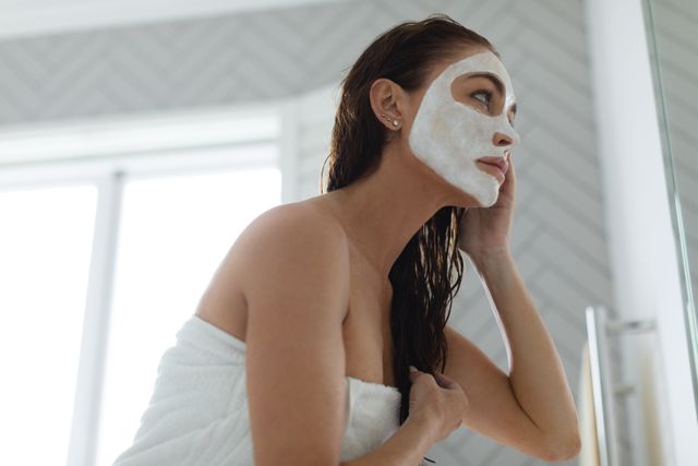 Woman wrapped in towel applying facial mask while looking in mirror in bathroom. Perfect for articles on skincare routines, beauty tips, self-care practices, spa treatments, and wellness lifestyle.