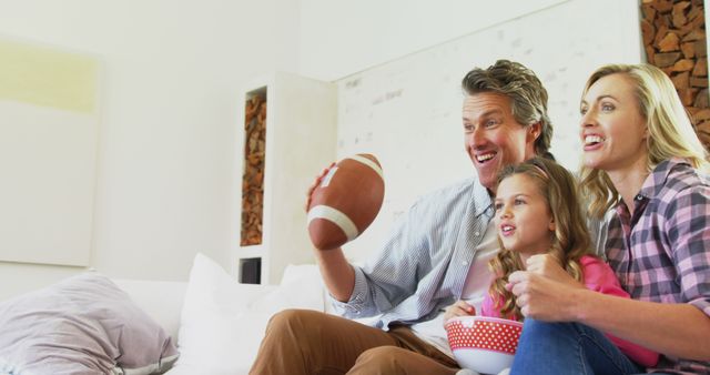 Parents and daughter enjoying American football game in living room. They hold a football and a bowl of snacks, smiling and looking excited. Ideal for themes of family bonding, leisure activities, sports enthusiasm, and cozy home lifestyle.