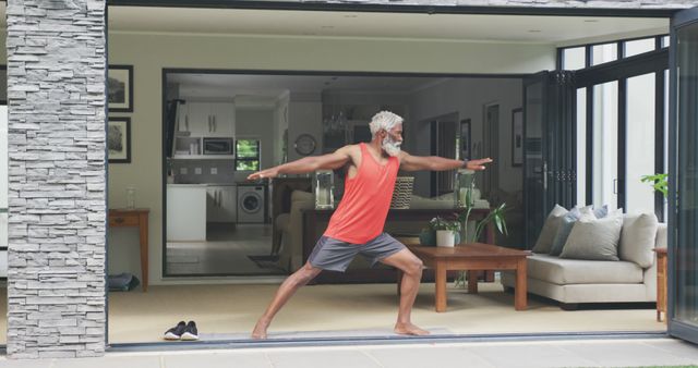 Senior man practicing yoga indoors in living room. Wearing casual exercise clothing in spacious modern home. Promotes concepts related to active aging, home-based exercise routines, maintaining physical fitness and overall well-being.