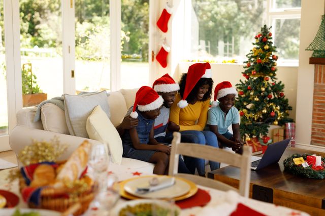 African-American family wearing Santa hats sitting on couch, engaging in a video call on laptop. Christmas tree and festive decorations in background. Christmas dinner table with bread and empty plate in foreground. Ideal for holiday greeting cards, family celebration promotions, and festive season advertisements.