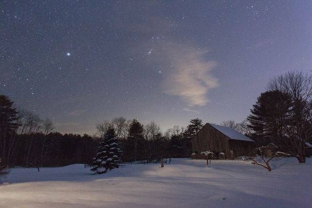 Picturesque night scene capturing an old rustic barn surrounded by a snow-covered landscape under a clear, starry sky. Suitable for winter travel guides, holiday cards, nature magazines, or digital wallpapers emphasizing tranquility, rural beauty, and winter's serene ambiance.