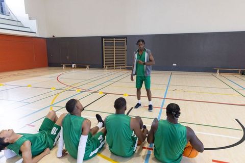 Coach coaching diverse male basketball team sitting and listening during indoor practice