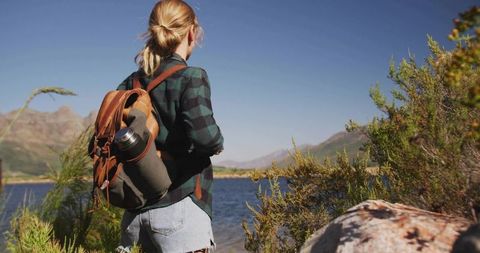 Young woman gazing toward distant mountain peaks from lakeshore with backpack