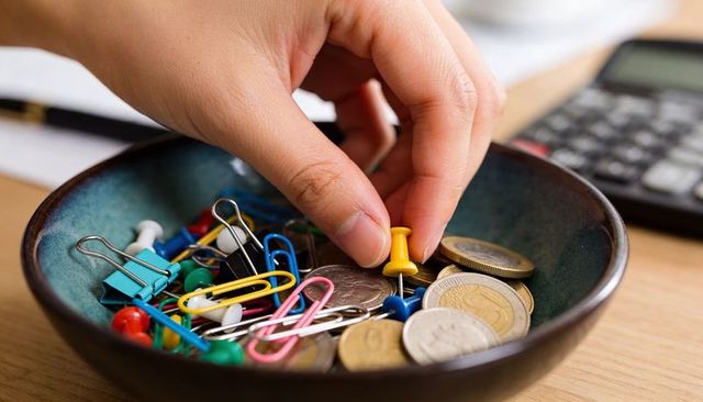 Hand picking yellow push pin from ceramic bowl of coins, paperclips and colorful office clips on des