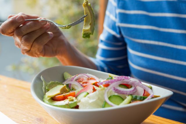 Senior woman enjoying a fresh vegetable salad with avocado, cucumber, tomato, and onion. Ideal for use in articles or advertisements about healthy eating, senior nutrition, wellness, and lifestyle choices. Perfect for promoting dietary tips, healthy recipes, and balanced meals.