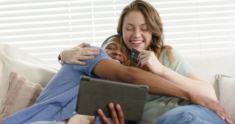 Couple cuddling and laughing on sofa holding tablet and credit card for online shopping