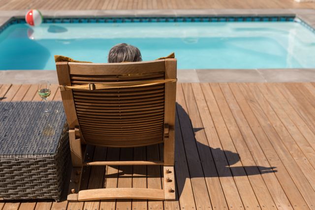 Senior African American woman enjoying a peaceful moment on a sun lounger by the swimming pool in her backyard. Ideal for use in lifestyle, retirement, and leisure-related content. Perfect for illustrating concepts of relaxation, summer activities, and outdoor living.