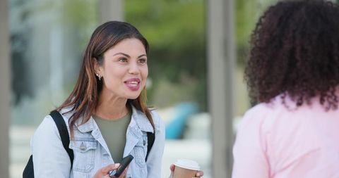 Female friends chatting on campus holding coffee and phone in casual denim jacket