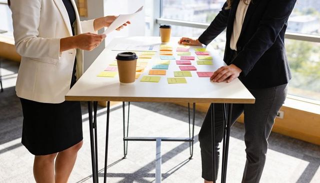 Businesswomen collaborating over sticky notes and coffee at tall table in bright office