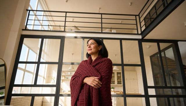 Middle-aged woman standing wrapped in maroon shawl gazing through sunlit loft windows