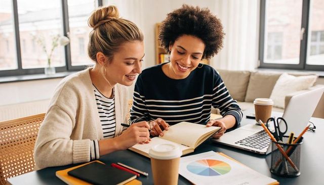 White and black women collaborating at sunlit home workspace writing notes with laptop
