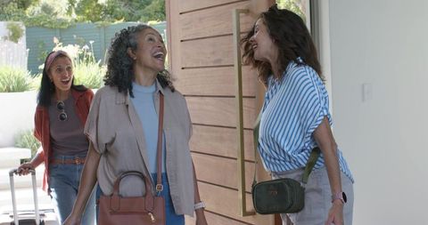 Three women reuniting at home entrance laughing, greeting returning traveler with luggage