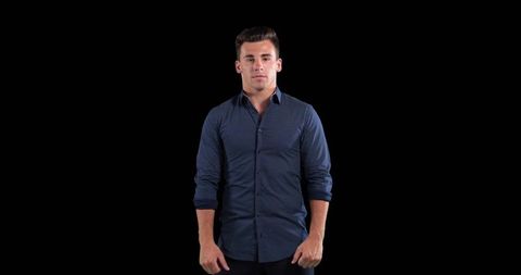 Confident young man standing wearing navy shirt and dark trousers against black background