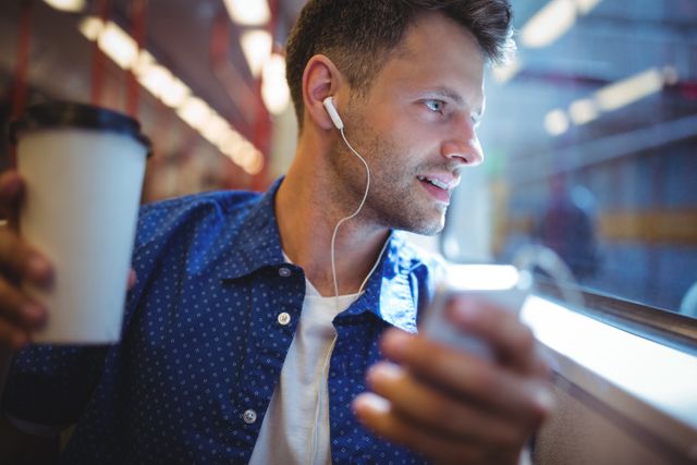 Man sitting on train, listening to music with headphones, holding coffee cup, looking out window. Ideal for use in travel blogs, public transport promotions, lifestyle articles, and advertisements related to commuting or urban living.