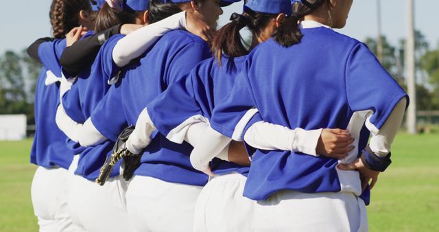 Female baseball players wearing blue uniforms huddling closely with arms around each other before a game. Ideal for topics on sportsmanship, teamwork, female empowerment in sports, and team unity.