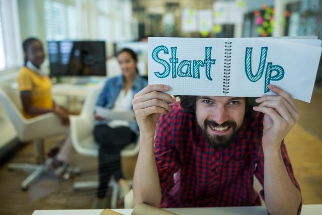Young entrepreneur smiling and holding a 'Start Up' sign in a modern office environment. Ideal for use in articles, blogs, and marketing materials related to entrepreneurship, business startups, innovation, and teamwork. Perfect for illustrating concepts of business growth, creative workspaces, and young professionals collaborating.