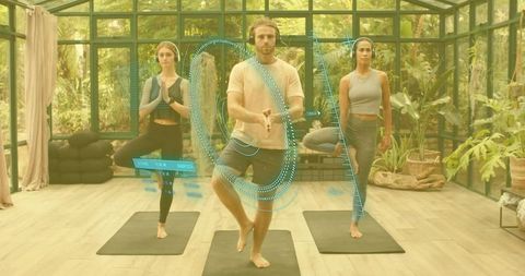 Three adults practicing ar-guided yoga tree pose in sunlit glasshouse studio