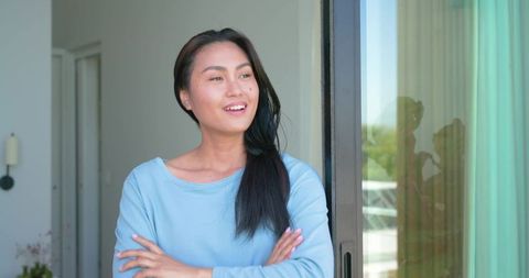 Mid-adult asian woman standing on balcony with arms crossed looking toward daylight reflection