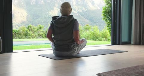 Senior asian man meditating on yoga mat at home with scenic mountain view