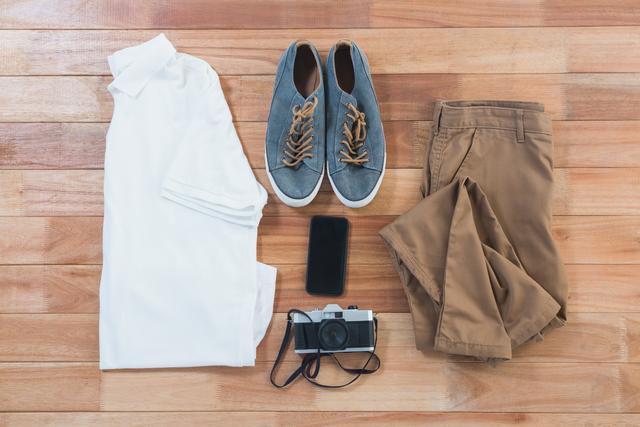 Flat lay of casual beach outfit and accessories including white shirt, blue shoes, brown shorts, vintage camera, and smartphone on wooden floor. Ideal for fashion blogs, travel websites, and lifestyle magazines.