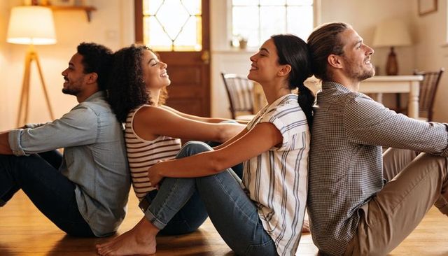Young diverse friends sitting back-to-back on hardwood floor in cozy living room