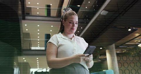 Young woman checking smartphone in modern coworking atrium with mezzanine and teal chair