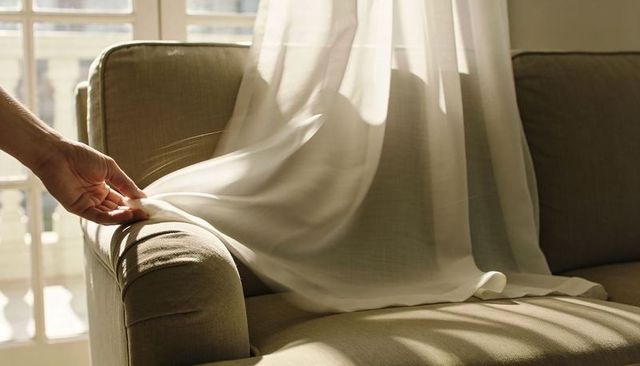 Hand smoothing sheer curtain over sunlit beige sofa, soft shadow play, minimalist interior