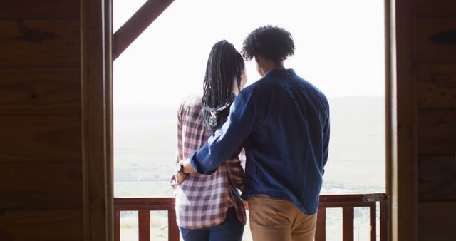 Happy african american couple looking through window and embracing in log cabin, slow motion. Lifestyle, domestic life, countryside and nature concept.