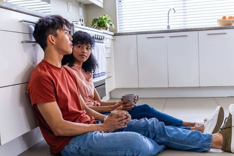 Young couple sitting on kitchen floor holding coffee mugs leaning on cabinets in sunlight