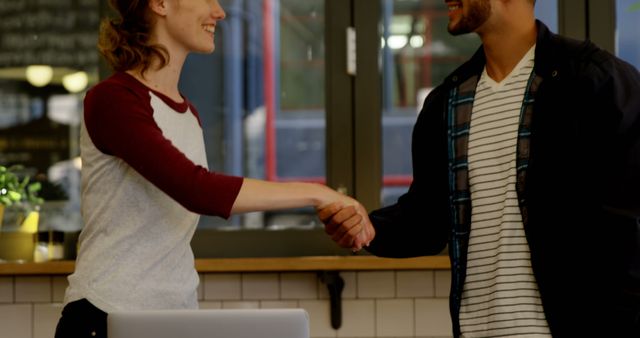A young Caucasian woman and a young man of Middle Eastern descent are shaking hands in a friendly manner in an indoor setting, with copy space. Their smiles suggest a positive interaction, indicating a successful meeting or agreement.
