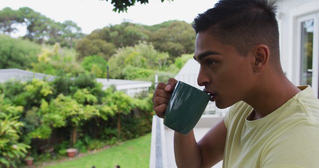 Biracial man standing on balcony drinking a mug of coffee looking at garden. staying at home in isolation during quarantine lockdown.
