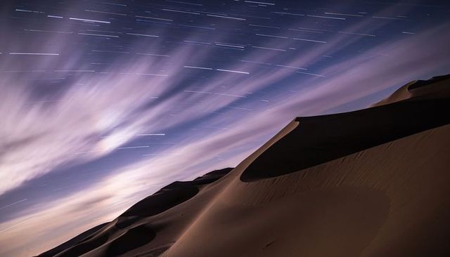Rising sand dune ridge under star trails and streaked clouds, long-exposure nightscape