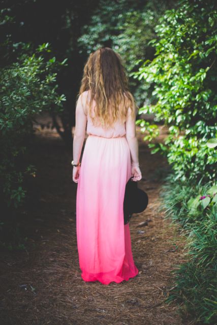 A woman with long, flowing hair is walking through a lush, green forest wearing a gradient pink dress. This image captures a serene and tranquil moment in a natural and vibrant environment. Excellent for use in themes of nature, tranquility, summer fashion, and outdoor adventures.
