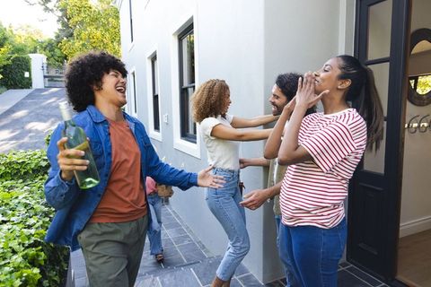 Diverse friends laughing and hugging at front porch holding wine bottle, celebrating