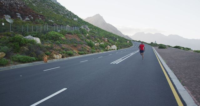 Man jogging on a scenic mountain road in the early morning light. He wears casual running attire. Ideal for advertising outdoor fitness regimes, healthy lifestyle choices, athletic gear, and travel promotions focusing on nature and adventure. Suitable for articles related to marathon training, wellness, and personal achievements.