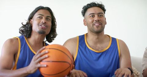 Two african american teammates sitting on couch holding basketball watching game